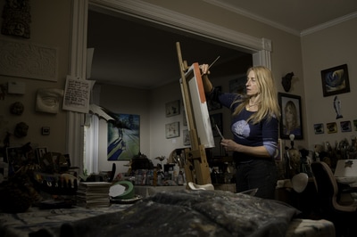 Artists Victoria Wallace gets back at a canvas that she had tucked away for a bit. Her work space is cluttered with tools, work to be done and completed paintings. The photographer, Mike Taylor, chose to light the scene very selectively.