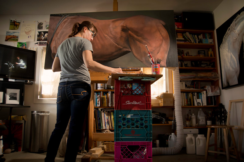 A woman applies paint to a large painting of a horse's body. The viewer is looking up from the floor.