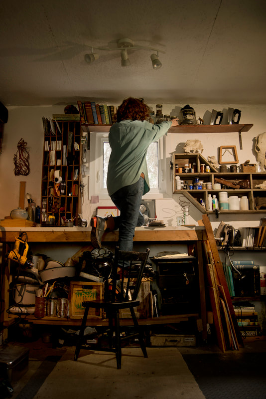 A woman climbs up on her workbench to reach some distant tools near the ceiling.