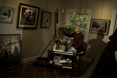 Doug Andrews painting in his home studio. His room walls are lined with framed artwork that he has created. His red sweater is dimly lit in this serene portrait by photographer Mike Taylor.
