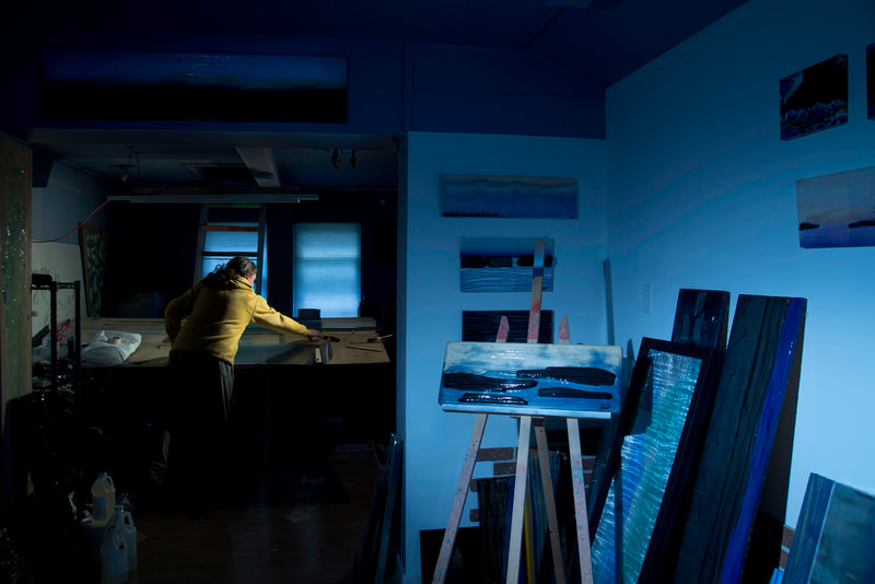 A man in a yellow sweater reaches for his work piece, the workshop is deeply blue in colour.