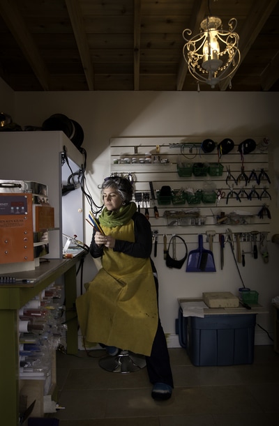 Annie Scherz sits in her small studio looking at her supplies. She wears a yellow leather apron that hangs down to her knees. The photographer Mike Taylor of Peterborough captured this image.