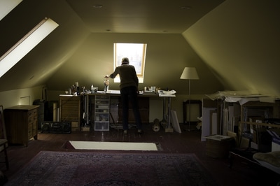 Artist Nan Sidler reaches for a paint brush as she is surrounded by gentle window light in her attic studio. The image is fairly dim, but yellow walls and ceiling are discernable in this portrait by photographer Mike Taylor of Peterborough.