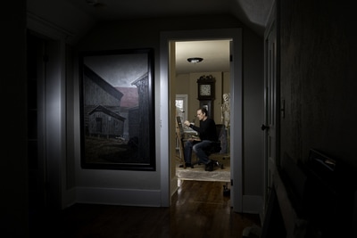 Patrick Fitzgerald who is both an illustrator and painter, takes his time to get the details right. His home studio is dimly lit, yet reveals a modeling skeleton behind him. Outside the room is a large painting, gently lit with a small light. Image by Peterborough photographer, Mike Taylor.