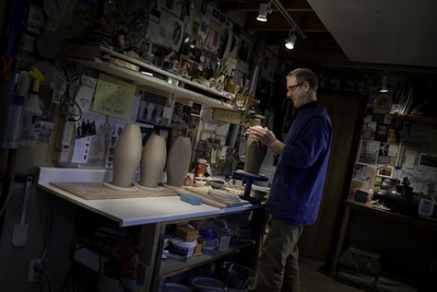 Thomas Aitken finishes shaping a tall vase in his basement pottery studio. The walls are filled with inspirational notes, photos, and various ephemera. The room is spot-lit to bring out the important bits in this Mike Taylor portrait.