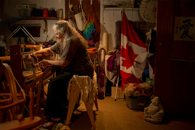A silver haired woman sits at a small loom, spinning yarn. A wrinkled Canadian flag rests in a distant corner.