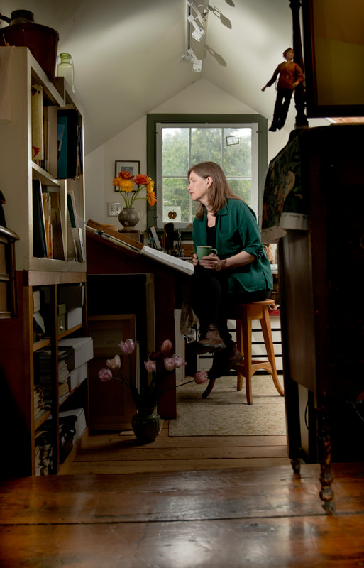 A woman in green top, sits on a stool in front of a window. She examines her art work on an easle.