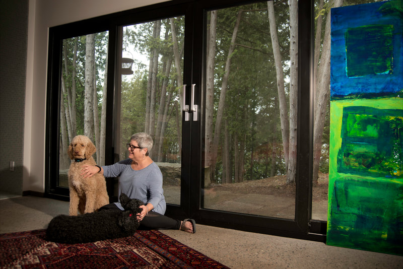 A woman sits on the carpet next to the glass doors. She is comforting her two poodles, one tan and the other black in colour.