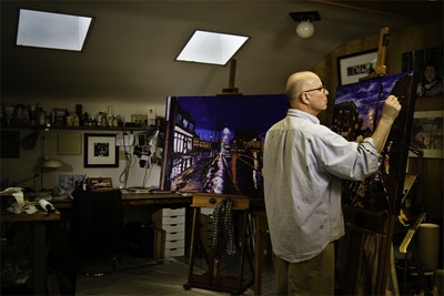 Rob Niezen is well known for his colourful work. Here he pays close attention to two new works. This portrait shows two colourful paintings, each on their own easel. Rob uses his right hand to apply finishing touches to the nearest of the paintings in this dimly lit space. Portrait number one, in the series by photographer Mike Taylor.
